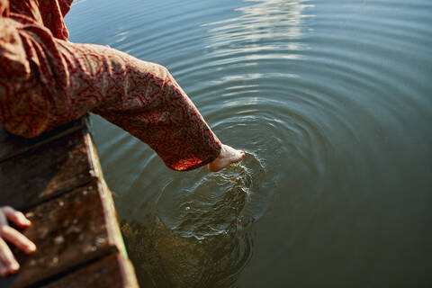 Woman sitting on jetty at a lake touching the water with her foot stock photo