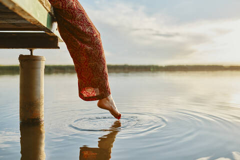 Frau sitzt auf einem Steg an einem See bei Sonnenuntergang und berührt das Wasser mit ihrem Fuß, lizenzfreies Stockfoto