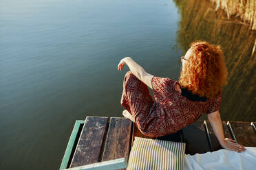 Redheaded young woman sitting on jetty at a lake at sunset - ZEDF03607