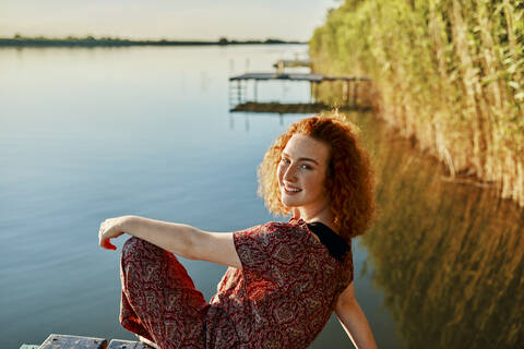 Porträt einer lächelnden rothaarigen jungen Frau, die auf einem Steg an einem See bei Sonnenuntergang sitzt, lizenzfreies Stockfoto