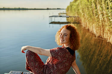 Portrait of smiling redheaded young woman sitting on jetty at a lake at sunset - ZEDF03608