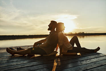 Couple sitting back to back on jetty at a lake at sunset - ZEDF03611