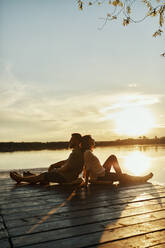 Paar sitzt Rücken an Rücken auf einem Steg an einem See bei Sonnenuntergang - ZEDF03612