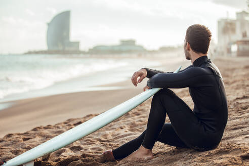 Seitenansicht eines Mannes in einem schwarzen Neoprenanzug, der mit einem Surfbrett am Sandstrand sitzt und nachdenklich in die Ferne schaut, Spanien - ADSF00097