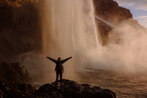 Rückenansicht eines Touristen, der mit gespreizten Händen an einem Wasserfall in Island steht. - ADSF00104