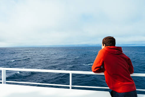 Back view of child in red hoodie standing on ship deck looking at endless blue sea expanses - ADSF00115