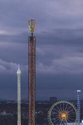 Germany, Bavaria, Munich, Aerial view of illuminated free fall and chain swing towers at dusk - MMAF01353