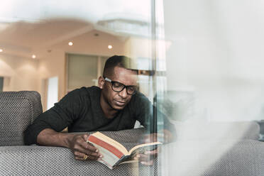 Adult African American man in glasses reading book while sitting on sofa at home - ADSF00127