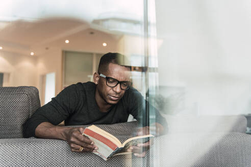 Erwachsener afroamerikanischer Mann mit Brille liest ein Buch, während er zu Hause auf dem Sofa sitzt - ADSF00127