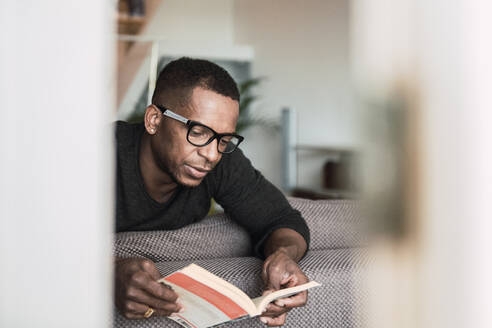 Erwachsener afroamerikanischer Mann mit Brille liest ein Buch, während er zu Hause auf dem Sofa sitzt - ADSF00128