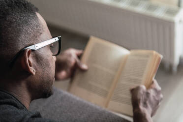 Erwachsener afroamerikanischer Mann mit Brille liest ein Buch, während er zu Hause auf dem Sofa sitzt - ADSF00129