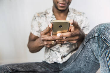 Closeup of shiny modern mobile phone in hands of black man sitting against white wall - ADSF00130