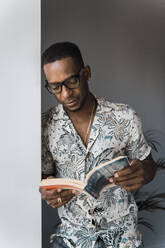 Casual black man in eyeglasses leaning on white wall near window and reading book - ADSF00131