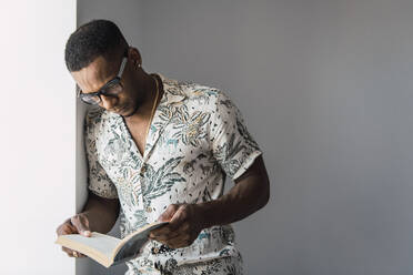 Lässiger schwarzer Mann mit Brille, der an einer weißen Wand in der Nähe des Fensters lehnt und ein Buch liest - ADSF00132