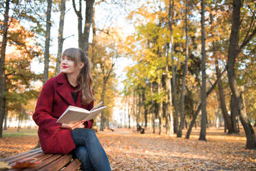 Attractive young�lady in red coat holding opened volume, sitting on seat and looking away in autumn forest - ADSF00142
