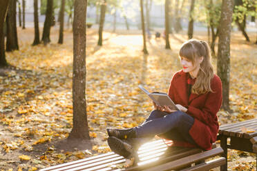 Attraktive junge Frau in rotem Mantel, die ein aufgeschlagenes Buch hält und auf einem Sitz im Herbstwald sitzt - ADSF00148