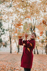 Stilvolle Frau in rotem Mantel wirft bunte gefallene Blätter im Park auf und lacht - ADSF00151