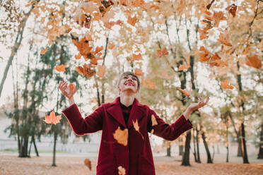 Stylish woman in red coat throwing up colorful fallen leaves in park and laughing - ADSF00152