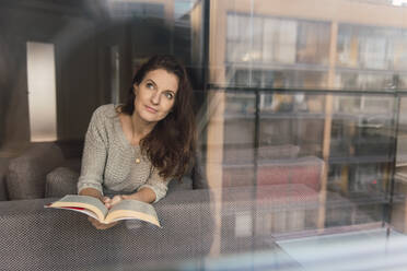 Lovely adult lady in elegant outfit smiling and looking away while lying on comfortable sofa with interesting book - ADSF00154