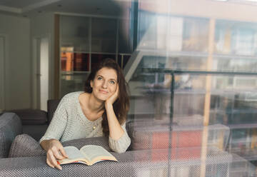 Schöne erwachsene Dame in elegantem Outfit lächelnd und wegschauen, während auf bequemen Sofa mit interessanten Buch liegen - ADSF00156