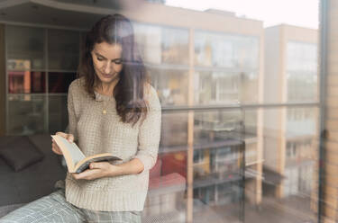 Erwachsene Frau mit Buch steht in der Nähe von Sofa und Fenster - ADSF00157