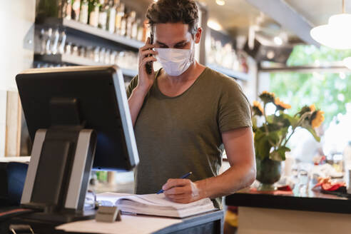 Restaurantleiter mit Schutzmaske, der einen Computer und ein Smartphone für die Reservierung benutzt - DIGF12752