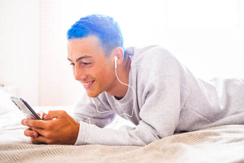 Junger Mann mit blauen Haaren, der auf dem Bett liegend sein Smartphone benutzt und Musik hört - SIPF02181