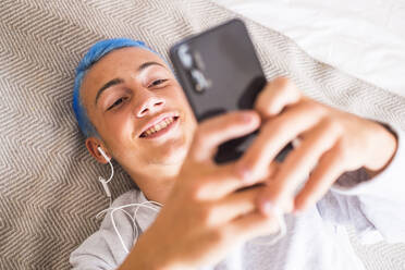 Junger Mann mit blauen Haaren, der auf dem Bett liegend sein Smartphone benutzt und Musik hört - SIPF02182