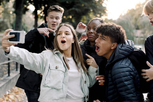 Cheerful teenage friends taking selfie through mobile phone while standing on street in city - MASF18646