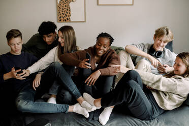 Lächelnde Freunde im Teenageralter, die soziale Medien auf ihren Handys nutzen, während sie sich zu Hause auf dem Sofa entspannen - MASF18664