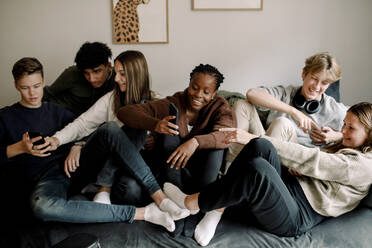 Smiling teenage friends using social media on mobile phones while relaxing on sofa at home - MASF18664