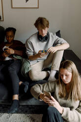 Teenage girls and boy using mobile phone while sitting in living room - MASF18668