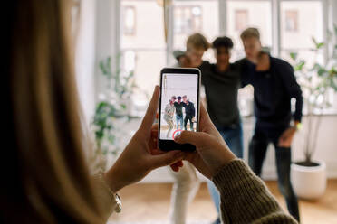 Teenage girl with smart phone filming male friends standing arm around in living room - MASF18674