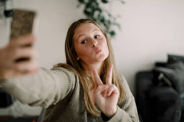Teenager-Mädchen zeigt Friedenszeichen bei der Aufnahme eines Selfies mit dem Handy zu Hause - MASF18686