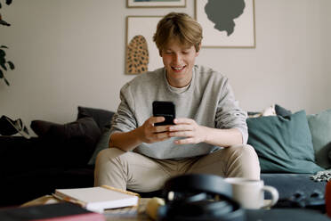 Happy teenage boy using mobile phone while sitting on sofa at home - MASF18689