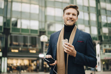 Portrait of smiling businessman with wrap sandwich standing in city - MASF18705