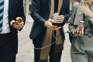 Midsection von Geschäftsleuten, die eine Brezel und ein Getränk halten, während sie ein Smartphone in der Stadt benutzen - MASF18706