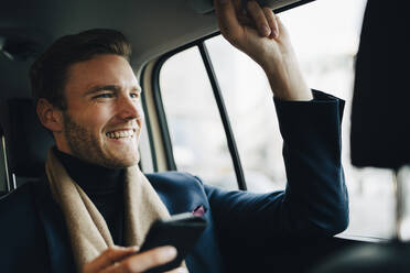 Lächelnder Geschäftsmann mit Telefon, der wegschaut, während er im Taxi sitzt - MASF18712