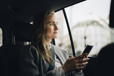 Nachdenkliche Unternehmerin mit Smartphone, die durch das Fenster schaut, während sie im Taxi sitzt - MASF18714