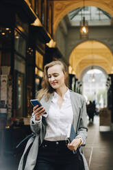 Smiling businesswoman using smart phone while standing in city - MASF18734