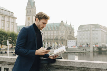 Geschäftsmann mit Smartphone schaut in ein Buch, während er in der Stadt steht - MASF18737