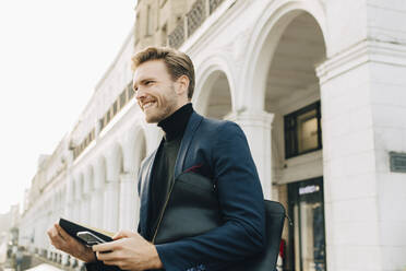 Lächelnder Geschäftsmann mit Smartphone, der wegschaut, während er an einem Gebäude in der Stadt steht - MASF18739
