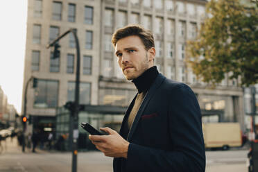 Nachdenklicher Geschäftsmann mit Smartphone, der wegschaut, während er an einem Gebäude in der Stadt steht - MASF18746
