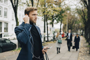 Businessman looking away while holding in-ear headphones in city - MASF18758