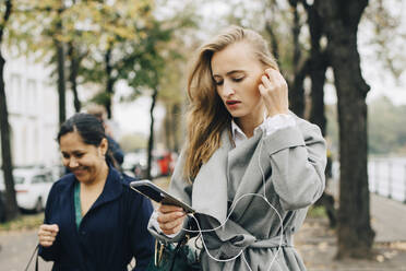 Geschäftsfrau trägt In-Ear-Kopfhörer und schaut in der Stadt auf ihr Smartphone - MASF18759