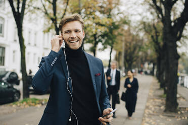 Smiling businessman looking away while holding in-ear headphones in city - MASF18760