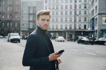 Side view of entrepreneur with phone standing against building in city - MASF18770