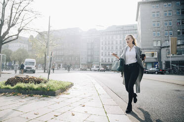 Unternehmerin in voller Länge mit Smartphone beim Spaziergang in der Stadt - MASF18774