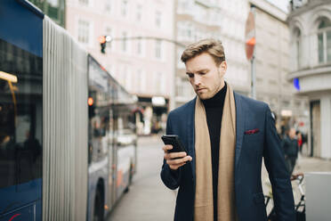 Männlicher Unternehmer, der das Internet über sein Mobiltelefon nutzt, während er in der Stadt am Bus steht - MASF18783