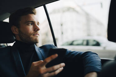 Männlicher Unternehmer mit Telefon, der durch das Fenster schaut, während er im Taxi sitzt - MASF18790